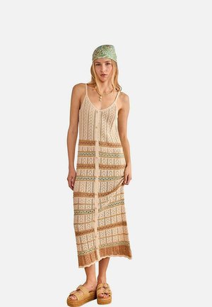 Mujer con vestido de punto sin mangas, con botones al frente y estampado, pañuelo verde en la cabeza, collares en capas y sandalias de plataforma.