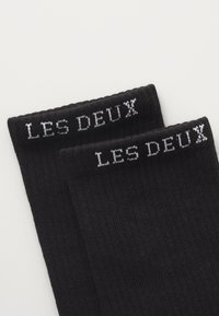 Sorte ribbede sokker med teksten "LES DEUX" i kontrastfarve hvid ved kanten, lavet af et blødt, fleksibelt materiale.