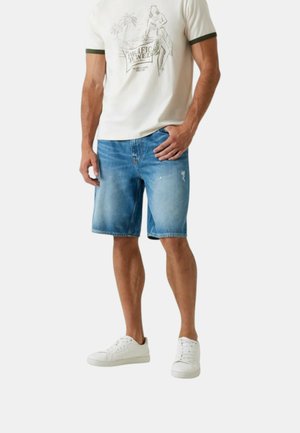 Uomo con maglietta bianca con grafica, pantaloncini di jeans blu leggermente consumati e scarpe basse bianche, in piedi con una mano in tasca.