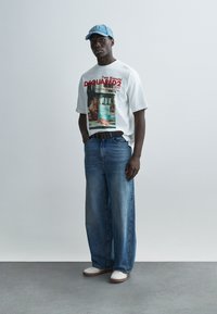 Λευκό γραφικό t-shirt με φωτογραφικό τύπωμα και κόκκινο κείμενο, συνδυασμένο με άνετα μπλε τζιν παντελόνια και ανοιχτό γαλάζιο καπέλο. Λευκά αθλητικά παπούτσια.