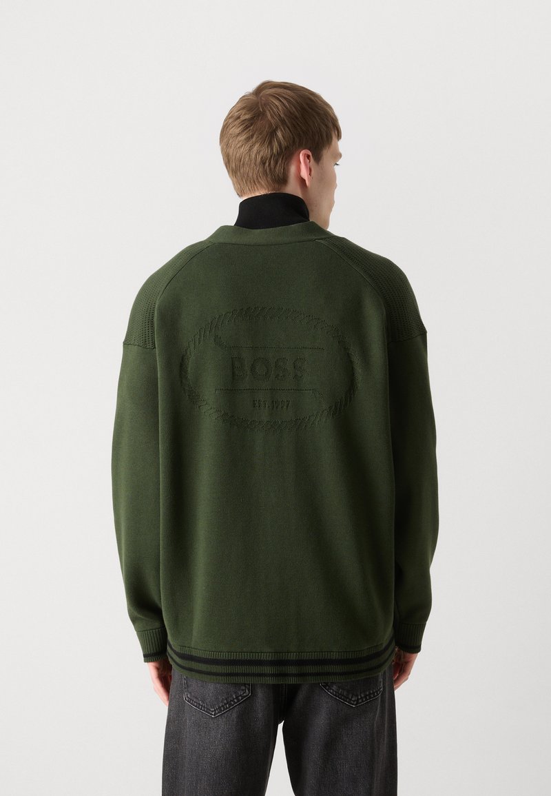 Zielony sweter dzianinowy z czarnym kołnierzem, z wytłoczonym logo "BOSS" na plecach. Żebrowane mankiety i dół dodają tekstury.