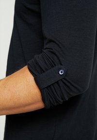 Camisa negra de manga larga con diseño de mangas fruncidas y detalle de botón. Fabricada en una tela suave y texturizada.