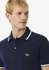 Polo de color azul marino hecho de tela texturizada, con un cuello plano a rayas azules, una tapeta de tres botones y un pequeño logo verde en el pecho.