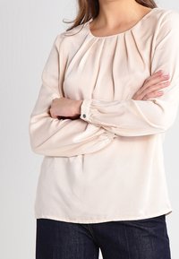 Blusa color crema con scollatura arricciata, maniche lunghe con polsini abbottonati e un tessuto liscio e leggero.