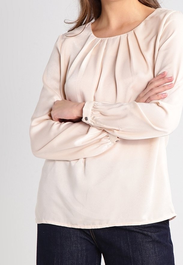 Blusa color crema con scollatura arricciata, maniche lunghe con polsini abbottonati e un tessuto liscio e leggero.