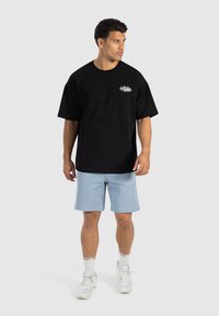Schwarzes Baumwoll-T-Shirt mit kurzen Ärmeln und einem runden weißen Logo auf der Brust, kombiniert mit hellblauen Shorts und weißen Turnschuhen.