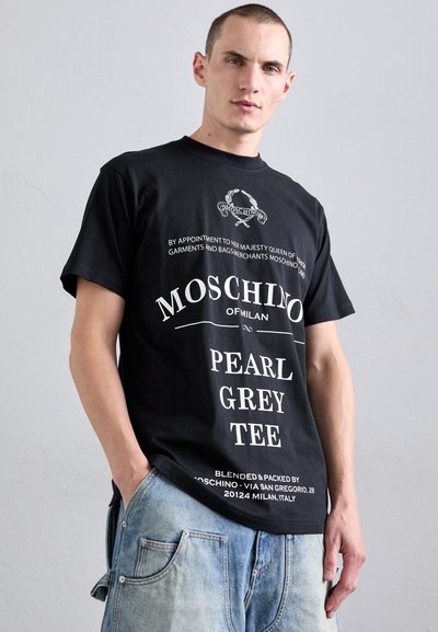 Juodas marškinėliai su balta tekstu, vaizduojantys "Moschino" prekės ženklą, "Pearl Grey Tee" ir dekoracijas. Minkšta medžiaga, standartinis kryžius.