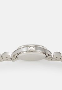 Rustfritt stålsur med en glatt, rund klokkehette, lenket armbånd og en sidekrone med logo. Minimalistisk design med polert overflate.