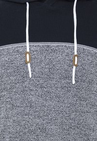 Gros plan d'un sweat à capuche en tricot avec une partie supérieure bleu marine, une partie inférieure gris chiné, des cordons blancs et des embouts de cordon marron.
