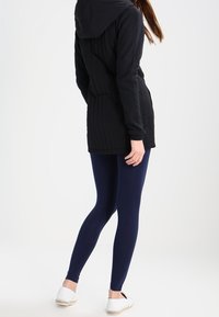 Chaqueta negra con capucha y costuras verticales, combinada con leggings azul marino. Alpargatas blancas con suelas de yute. Diseño simple y aerodinámico.