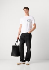 Hvid bomulds T-shirt med en frontlomme, der har et grafisk design, parret med mørke denim jeans og en sort læder skuldertaske.