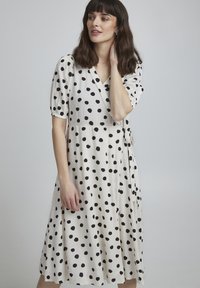 Vestido blanco cruzado con grandes lunares negros, mangas cortas con volumen y un lazo en la cintura, hecho de una tela ligera.