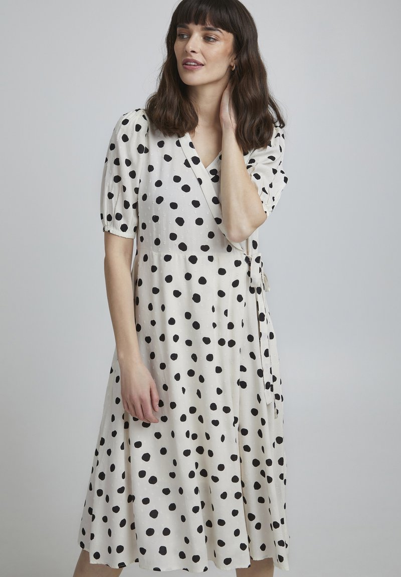 Vestido blanco cruzado con grandes lunares negros, mangas cortas con volumen y un lazo en la cintura, hecho de una tela ligera.