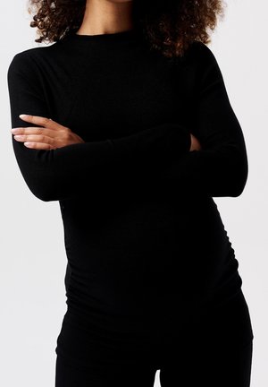 Woman with curly hair wearing a fitted black long-sleeve top, arms crossed, set against a plain light gray background.