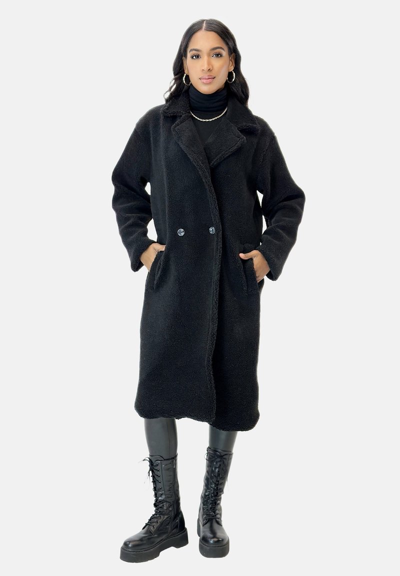 Svart shearling-kappa med urklippt krage, dubbla knappar och sidofickor; tillsammans med svarta läder boots med snörning.