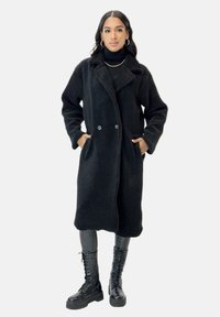 Schwarzer, langer Wollmantel mit strukturierter Oberfläche, doppelreihiger Vorderseite und Seitentaschen, kombiniert mit schwarzen Leggings und Kampfstiefeln.