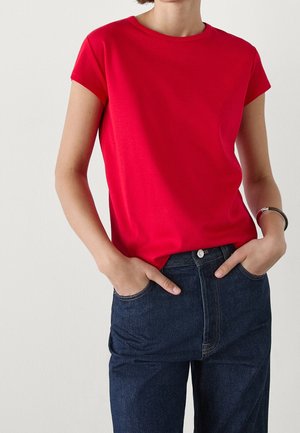 Kobieta w prostym czerwonym t-shircie z krótkim rękawem wpuszczonym w ciemnoniebieskie jeansy z wysokim stanem, z rękami w kieszeniach i srebrną bransoletką na lewej ręce.