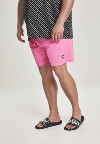 Pantaloni da bagno rosa con vita con coulisse e piccolo logo nero, abbinati a sandali neri con testo bianco. Camicia nera a fantasia sopra.