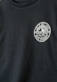 Melna T-kreklu ar baltu kalnu logotipu un tekstu "Iesim pazaudēties, mežonīga ceļojuma pārgājiens, paliec mežonīgs" uz krūtīm.