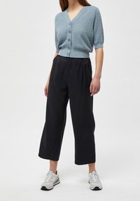 Světlomodrý pletený cardigan s výstřihem do V a zapínáním na knoflíky, doplněný černými zkrácenými kalhotami. Oblečený s bílými a šedými sportovními teniskami.