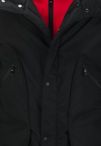 Schwarze gepolsterte Jacke mit rotem Innenfutter, ausgestattet mit Reißverschluss-Seitentaschen und Druckknopfverschlüssen. Robustes Gewebe mit strukturierter Oberfläche.
