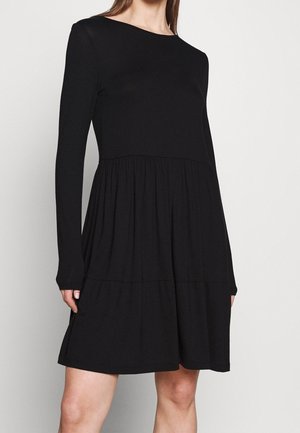 Frau trägt ein langärmeliges schwarzes Kleid mit einem lockeren, gerafften Rock und steht vor einem schlichten hellen Hintergrund.