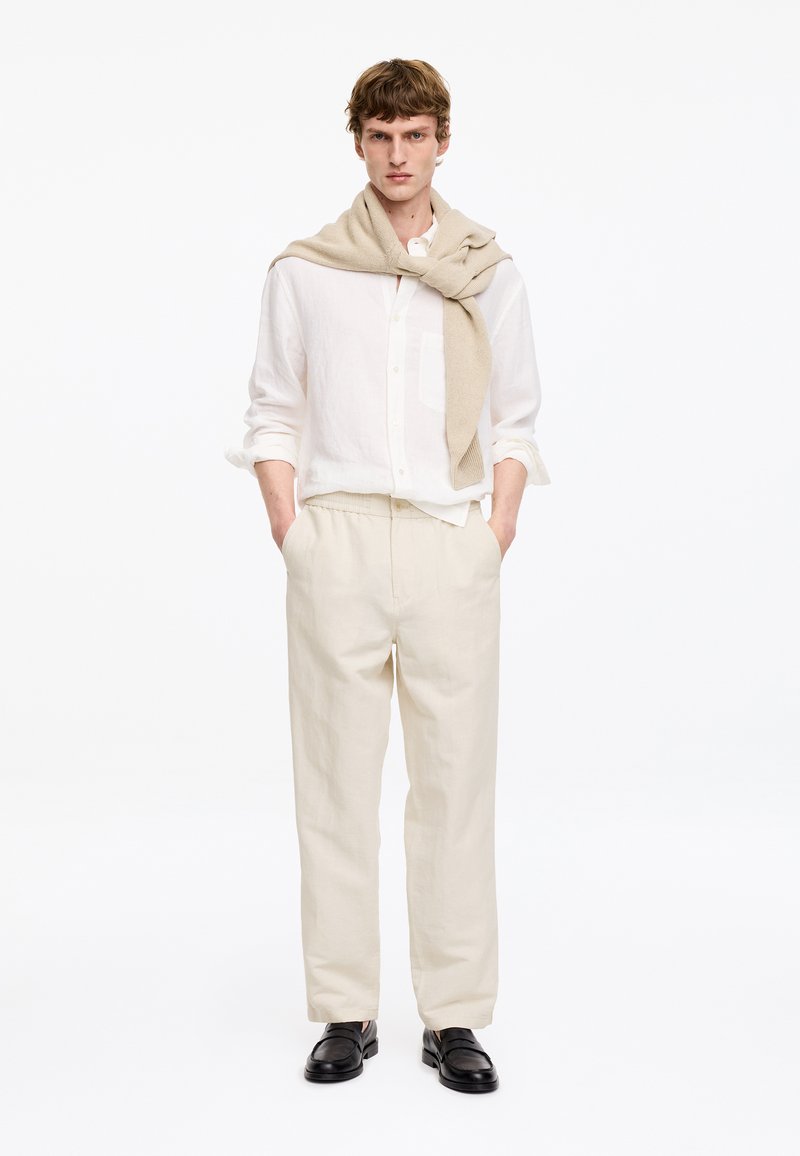 Jeune homme portant une chemise blanche boutonnée, un pantalon beige clair, des chaussures noires et un pull beige drapé sur les épaules.