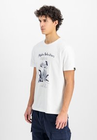 Tricou alb din bumbac cu un imprimeu grafic reprezentând o femeie ținând un tavă, cu textul "Alpha Industries" deasupra. Gât rotund, mâneci scurte.