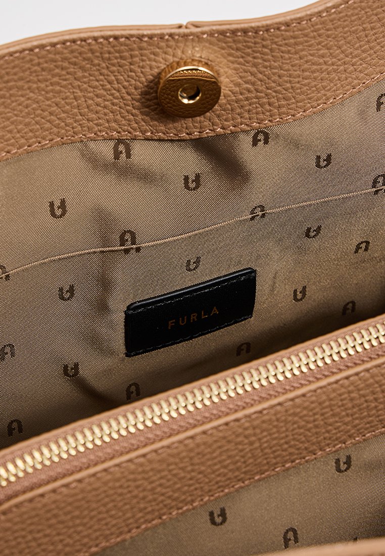 Innenraum einer handgefertigten Tasche aus braunem Leder mit beigem Stofffutter, das mit kleinen dunklen Logos gemustert ist, einem Reißverschlussfach und einem schwarzen Etikett mit der Aufschrift "Furla."