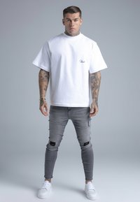 Fehér, túlméretezett póló kis logóval, világosszürke, karcsúsított farmerral, mely szakadt részleteket tartalmaz, és fehér tornacipővel párosítva.