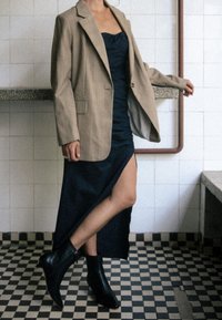 Brun rutig oversized blazer, svart draperad klänning med slits och spetsiga svarta ankelstövlar. Texturerade ytor och kontrasterande färger.