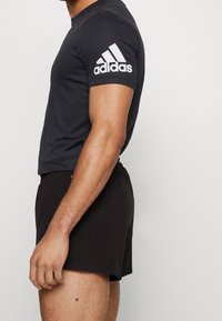 Svart atletisk t-shirt med vitt Adidas-logotyp på ärmen, matchad med svarta shorts med perforerad textur.