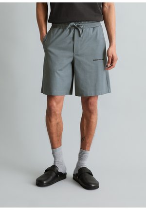 Man draagt grijze shorts met trekkoord en het logo "Marc O'Polo Denim", grijze sokken en zwarte instapschoenen, staand tegen een effen achtergrond.