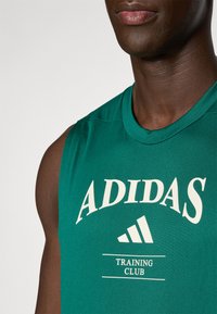 Zaļš bezpiedurkņu sporta krekls ar "ADIDAS" logo krēmkrāsā un tekstu "TRAINING CLUB" zem tā, izgatavots no gluda, viegla materiāla.