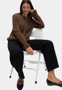 Brązowy sweter dzianinowy z perłowymi guzikami, prążkowanymi mankietami i falbaniastym dołem. Noszony na czarnych szerokich spodniach i czarnych baletkach.