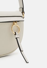 Bolso cruzado estructurado en material beige claro con ribetes negros, que presenta un acento de hardware circular en tono dorado y una correa plana para el hombro.