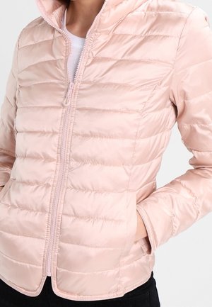 Person trägt eine hellrosa gesteppte Jacke mit Reißverschluss und seitlichen Taschen, die Hände lässig in den Taschen, über einem weißen Innenhemd.