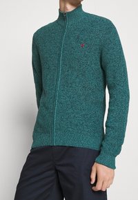 Turkos zip-up-tröja med hög krage och långa ärmar, med en strukturerad stickad mönster och en liten röd logga på bröstet.