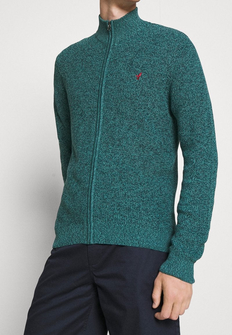 Turkos zip-up-tröja med hög krage och långa ärmar, med en strukturerad stickad mönster och en liten röd logga på bröstet.