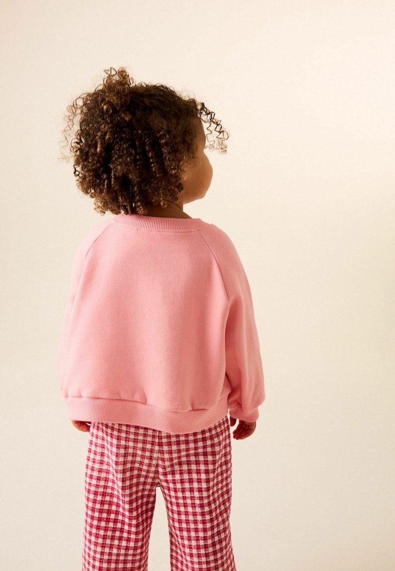 Felpa rosa con polsini a coste, abbinata a pantaloni a quadri rossi e bianchi. Il bambino ha capelli ricci ed è visto di spalle.