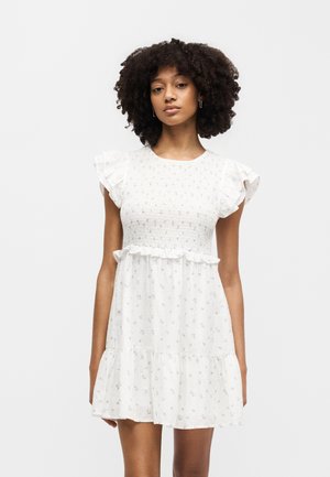 Mladá žena s kučeravými vlasmi v bielych krátkych kvetovaných šatách s volánikmi na rukávoch a spodnom leme, stojaca pred jednoduchým pozadím.