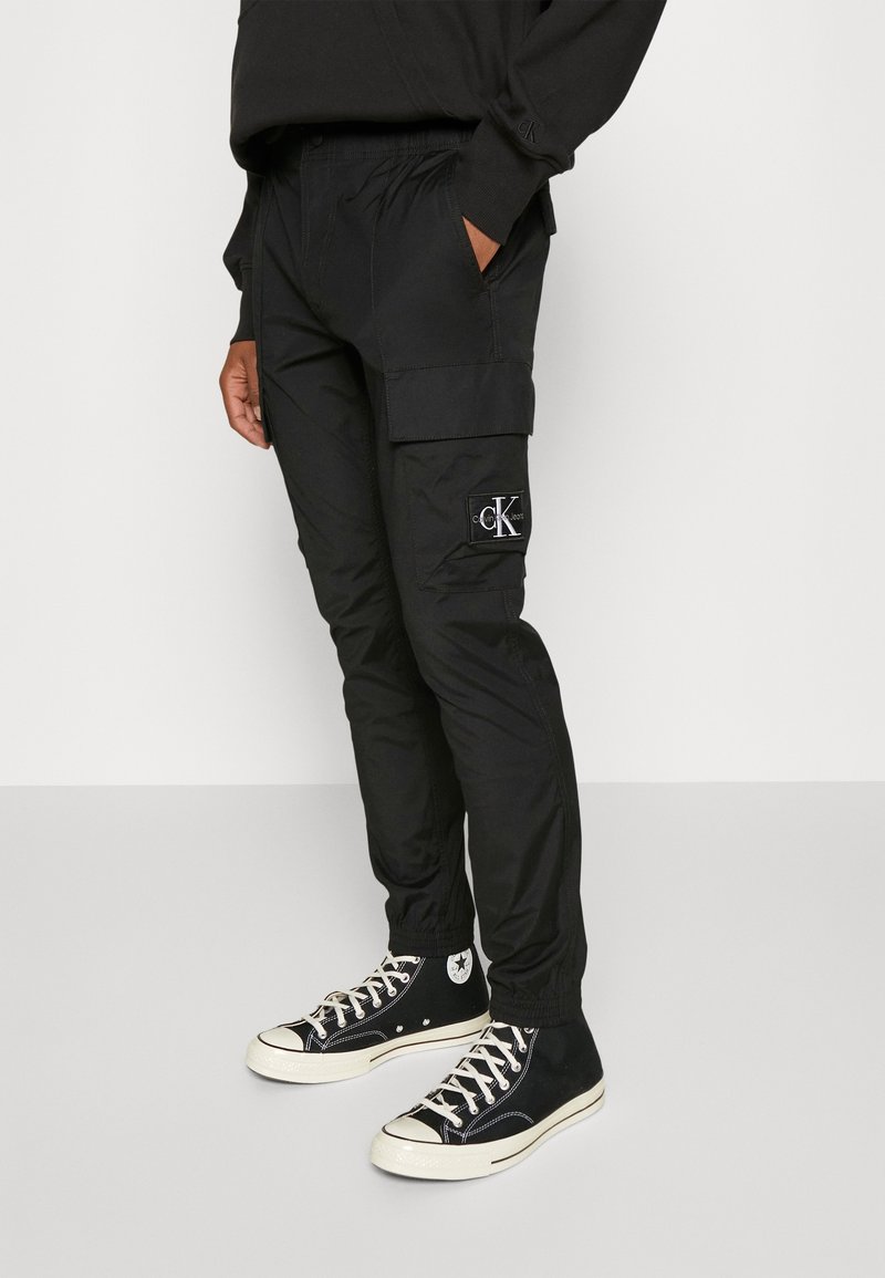 Svarta cargobyxor i lätt material, med sidofickor, elastiska muddar och en liten vit logotyp på låret.