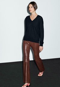 Donna che cammina con un maglione nero a scollo a V, pantaloni in pelle marrone e sandali rossi con cinturini, su uno sfondo chiaro e semplice.