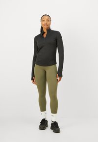 Un top negru cu mânecă lungă și fermoar la jumătate, asortat cu colanți verzi măslinii, adidași negri și șosete albe, având un design minimal și o formă ajustată.