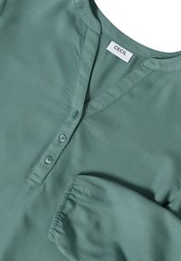 Grön långärmad blus med rund halsringning, tre framknappar, elastiska ärmslut och en "CECIL"-etikett inuti kragen.