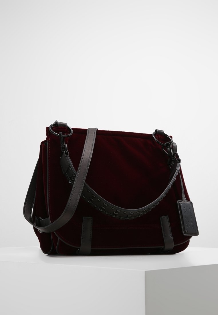 Bolso de terciopelo burdeos con una correa gris desmontable, detalles en negro y una etiqueta. Presenta una forma estructurada y costuras decorativas.