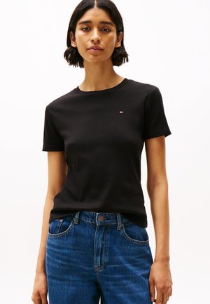 Rövid sötét hajú nő fekete kerek nyakú pólót visel, amelyen kis logó található a mellkasán, valamint kék farmernadrágot, és fehér háttér előtt áll.