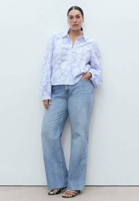 Chemise boutonnée violet clair avec une texture florale, associée à un jean léger et ample bleu clair et des sandales noires à lanières. Le modèle se tient dans un cadre neutre.