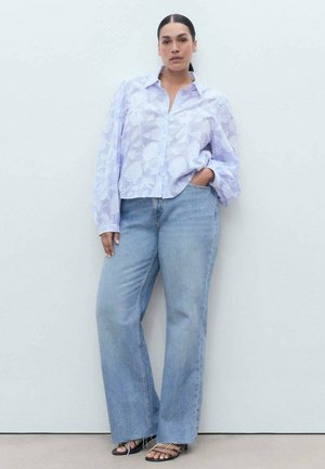 Helle lila Hemdbluse mit floralem Muster, kombiniert mit lockeren, hellblauen Jeans und schwarzen Sandalen mit Riemchen. Das Modell steht in einer neutralen Umgebung.