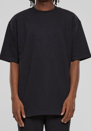 Person iført en løstsiddende, ensfarvet sort T-shirt med korte ærmer, vist fra skuldre til midt på låret mod en ensfarvet baggrund.